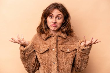 Beyaz tenli genç bir kadın bej arka planda izole edilmiş sorgulama sırasında omuz silkiyor ve şüphe duyuyor..