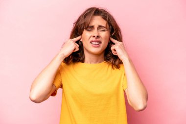 Pembe arka planda izole edilmiş genç beyaz bir kadın parmaklarıyla kulaklarını kapatıyor, gürültülü bir ortam yüzünden stresli ve çaresiz..
