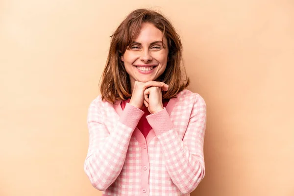 Young caucasian woman isolated on beige background keeps hands under chin, is looking happily aside.
