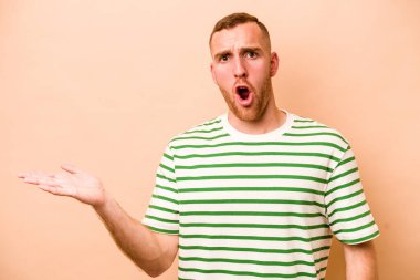 Beyaz tenli, bej arka planda izole edilmiş genç bir adam, avuç içi fotokopi alanı tutmaktan etkilenmiş..