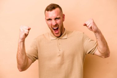 Beyaz tenli genç adam bej arka planda izole edilmiş tasasız ve heyecanlı tezahüratlar yapıyor. Zafer konsepti.