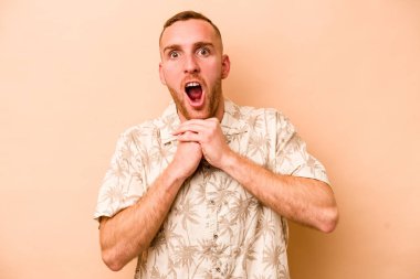 Beyaz tenli genç adam bej arka planda izole edilmiş şans için dua ediyor, şaşırmış ve ağzını açıp önüne bakıyor..