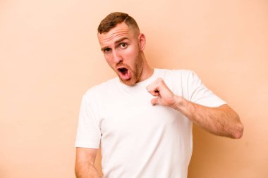 Beyaz tenli genç adam bej arka planda izole edildi. Parmakla göstererek şaşırdı, gülümsedi..