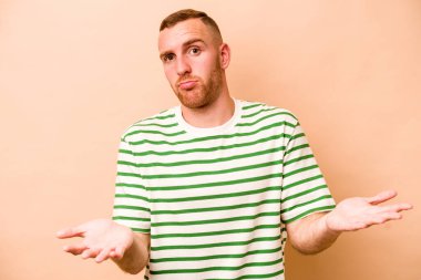 Beyazlı genç bir adam bej arka planda izole edilmiş sorgulama sırasında omuz silkiyor ve şüphe duyuyor..