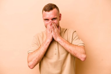 Beyazlı genç bir adam bej arka planda izole edilmiş bir şekilde bir şeye gülüyor, ağzı ellerle örtüyor..