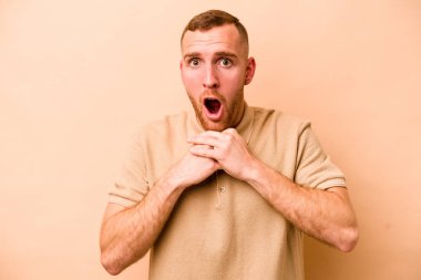 Beyaz tenli genç adam bej arka planda izole edilmiş şans için dua ediyor, şaşırmış ve ağzını açıp önüne bakıyor..
