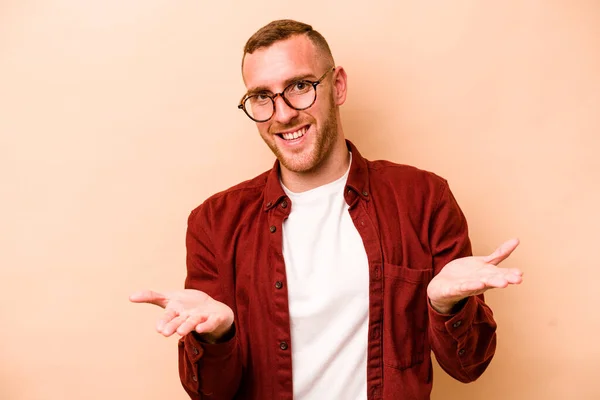 Young Caucasian Man Isolated Beige Background Makes Scale Arms Feels — Stock Photo, Image