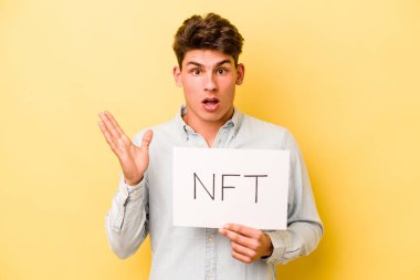 Sarı arka planda NFT pankartı tutan genç beyaz adam şaşırdı ve şok oldu..