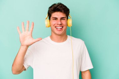 Mavi arka planda izole edilmiş müzik dinleyen beyaz bir adam gülümseyerek beş numarayı parmaklarıyla gösteriyor..