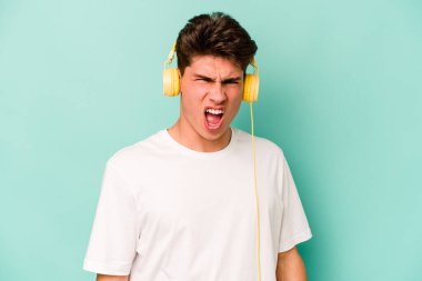 Mavi arka planda izole edilmiş müzik dinleyen genç beyaz adam çok sinirli ve agresif çığlıklar atıyor..