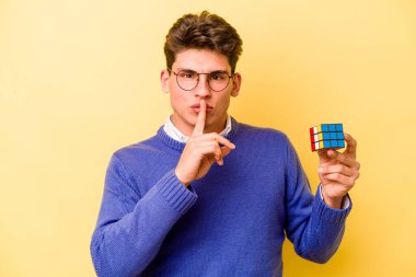 Sarı arka planda Rubiks küpü tutan genç beyaz bir adam bir sır saklıyor ya da sessizlik istiyor..
