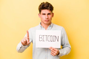Sarı arka planda Bitcoin levhası tutan genç beyaz adam parmak ile bir numarayı gösteriyor..