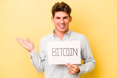 Sarı arka planda Bitcoin levhası tutan genç beyaz adam bir avuç içinde fotokopi alanı ve belinde bir el daha olduğunu gösteriyor..