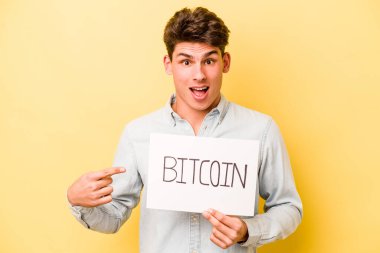 Sarı arka planda bitcoin levhası tutan genç beyaz adam yan tarafı işaret ediyor.