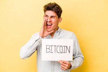 Sarı arka planda izole edilmiş Bitcoin levhası tutan genç beyaz adam bağırıyor ve avuç içini açık tutuyor..