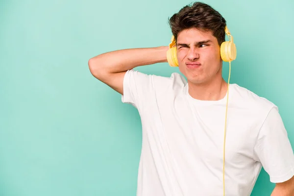 Young caucasian man listening to music isolated on blue background touching back of head, thinking and making a choice.