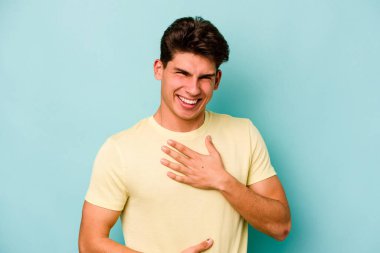 Mavi arka planda izole edilmiş genç beyaz adam mutlu bir şekilde gülüyor ve ellerini karnının üstünde tutarak eğleniyor..