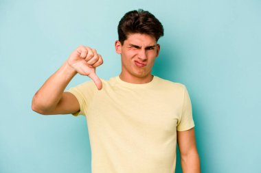 Mavi arka planda izole genç beyaz adam baş parmağını aşağı gösteriyor, hayal kırıklığı konsepti.