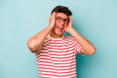 Mavi arka planda izole edilmiş genç beyaz adam ellerini başının üstünde tutarak neşe içinde gülüyor. Mutluluk kavramı.