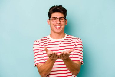 Kafkasyalı genç adam mavi arka planda izole edilmiş. Elinde avuç içi olan bir şey var ve kameraya sunuyor..