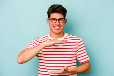 Mavi arka planda izole genç beyaz adam iki eliyle bir şey tutuyor, ürün sunumu..