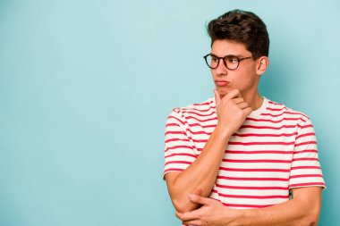 Mavi arka planda izole edilmiş genç beyaz adam şüpheli ve şüpheci bir ifadeyle yana bakıyor..