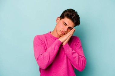 Mavi arka planda izole edilmiş genç beyaz adam esniyor. Ağzı eliyle kapatan yorgun bir hareket sergiliyor..