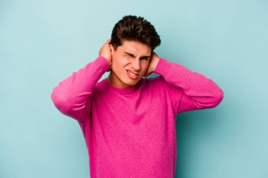 Mavi arka planda izole edilmiş genç beyaz adam kulaklarını elleriyle kapatıyor..