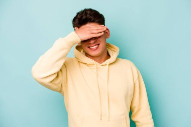 Mavi arka planda soyutlanmış genç beyaz bir adam elleriyle gözlerini kapatıyor, geniş bir gülümsemeyle bir sürpriz bekliyor..