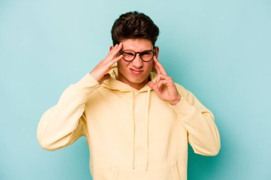 Genç beyaz adam mavi arka planda izole edilmiş tapınaklara dokunuyor ve başı ağrıyor..