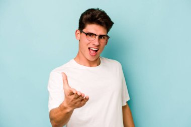 Mavi arka planda izole edilmiş genç beyaz adam selamlama jesti olarak kameraya elini uzatıyor..