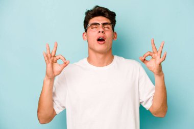 Mavi arka planda izole edilmiş genç beyaz adam yorucu bir günün ardından rahatlıyor. Yoga yapıyor..