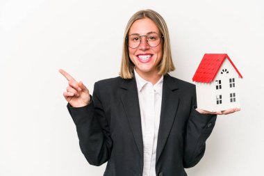 Beyaz arka planda izole edilmiş oyuncak evi tutan beyaz bir kadın gülümsüyor ve boş alanda bir şey gösteriyor..