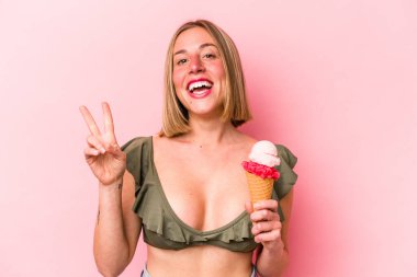 Bikini giyen beyaz bir kadın pembe arka planda izole edilmiş bir dondurma tutuyor neşeli ve kaygısız parmaklarıyla barış sembolü gösteriyor..