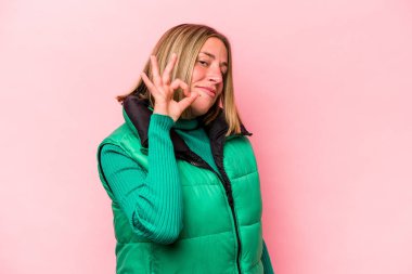 Pembe arka planda izole edilmiş genç beyaz kadın bir göz kırpıyor ve elinde iyi bir el hareketi tutuyor..