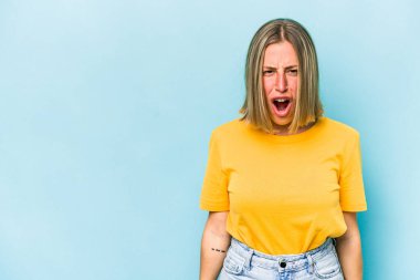 Kafkasyalı genç bir kadın mavi arka planda izole edilmiş çok kızgın, öfkeli, hayal kırıklığına uğramış bağırıyor..