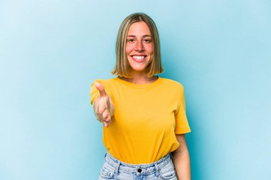 Mavi arka planda izole edilmiş genç beyaz kadın selamlama jesti olarak kameraya elini uzatıyor..