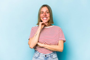 Mavi arka planda izole edilmiş genç beyaz kadın mutlu ve kendinden emin gülümsüyor, elleriyle çenesine dokunuyor..