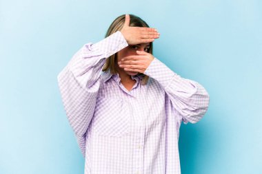 Mavi arka planda izole edilmiş genç beyaz kadın kameraya parmaklarıyla göz kırpıyor, yüzünü kapatmaktan utanıyor..