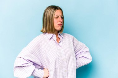 Mavi arka planda izole edilmiş genç beyaz kadın hoşnutsuzca kaşlarını çatmış, kollarını kavuşturmuş..
