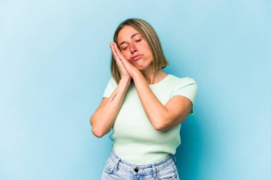 Mavi arka planda izole edilmiş genç beyaz kadın esniyor. Ağzı eliyle kapatan yorgun bir hareket sergiliyor..