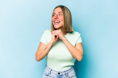 Mavi arka planda izole edilmiş genç beyaz kadın ellerini çenesinin altında tutuyor, mutlu bir şekilde yana bakıyor..