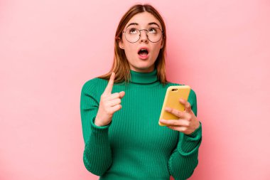 Elinde cep telefonu olan beyaz bir kadın pembe arka planda izole edilmiş ve ağzı açık bir şekilde üst tarafı işaret ediyor..