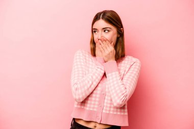 Pembe arka planda izole edilmiş genç beyaz kadın bir şeye gülüyor, elleriyle ağzını kapatıyor..