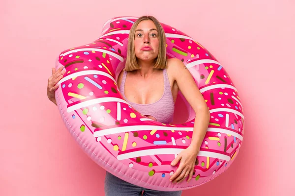 Beyaz bir kadın pembe arka planda izole edilmiş şişme bir yatak tutuyor omuzlarını silkiyor ve gözleri açık..