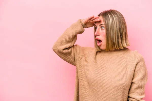 Pembe arka planda izole edilmiş genç beyaz kadın uzaklara bakıyor ve elini alnına koyuyor..