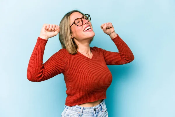 Mavi arka planda izole edilmiş genç beyaz kadın özel bir günü kutluyor, atlıyor ve enerjiyle kollarını kaldırıyor..