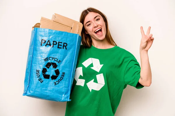 白地に隔離された紙をリサイクルするリサイクルバッグを持っている若い女性喜びと屈託のない指で平和のシンボルを示す — ストック写真