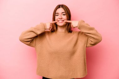 Pembe arka planda izole edilmiş genç beyaz kadın gülümsüyor, parmaklarını ağıza doğrultuyor..