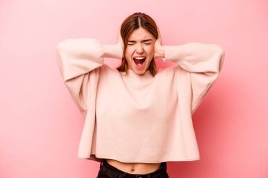 Pembe arka planda izole edilmiş genç, beyaz bir kadın. Kulakları kulaklarıyla kapatıyor. Çok fazla ses duymamaya çalışıyor..